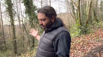 Man in the woods on a hill talking to camera. He is wearing a bodywarmer gilet and has long hair and beard.