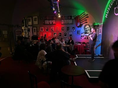 Louie Christie performing stand-up comedy while audience laughs. On the wall behind there is a big painting of Tommy
            Cooper and smaller photos of various famous stand up comedians. Louie is pointing at the audience and seems to be having
            a good time. His hair is in a man bun. He's pointing at a woman in the audience and she has her hands crossed in front of
            her face as if she doesn't want to talk. Which is ironic, because it was about the only time in the whole night that she
            shut up.