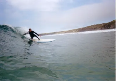 A surfer on a wave. He's crouching, touching the wave with his hand as the wave breaks. The wave is chest high,
            blue-green color. There are brown-green hills in the background on the land.